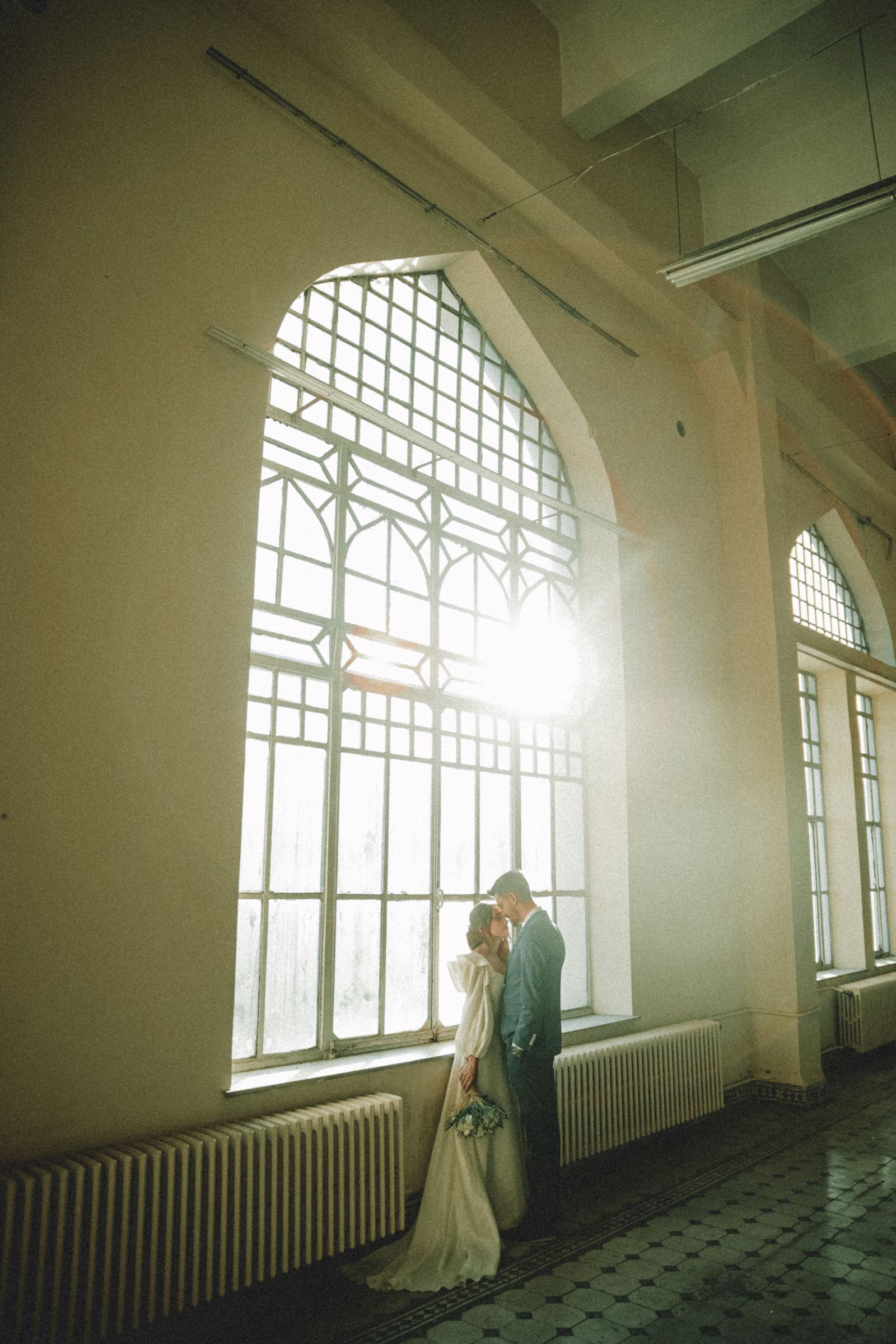 Bride and groom beneath grand arched windows in a sunlit historic hall